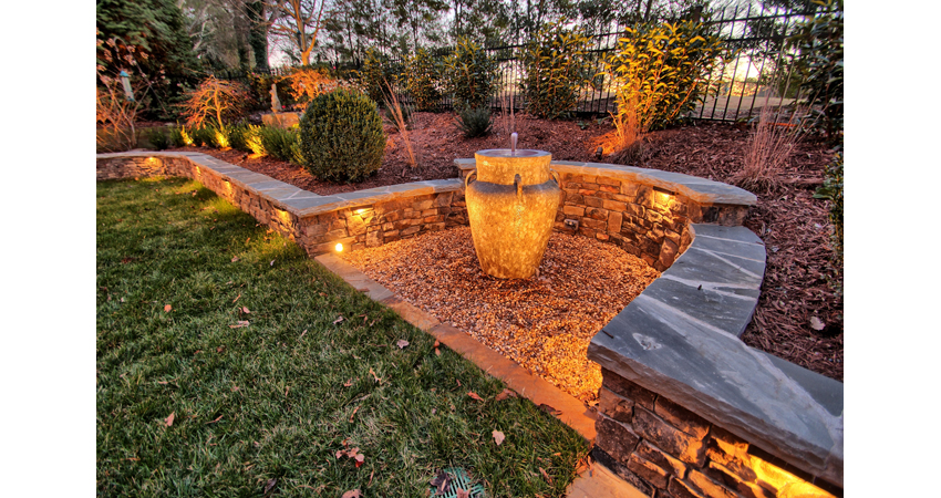 Backyard lighting along a stone wall and in the planting areas to add interest and light up a fountain at night.
