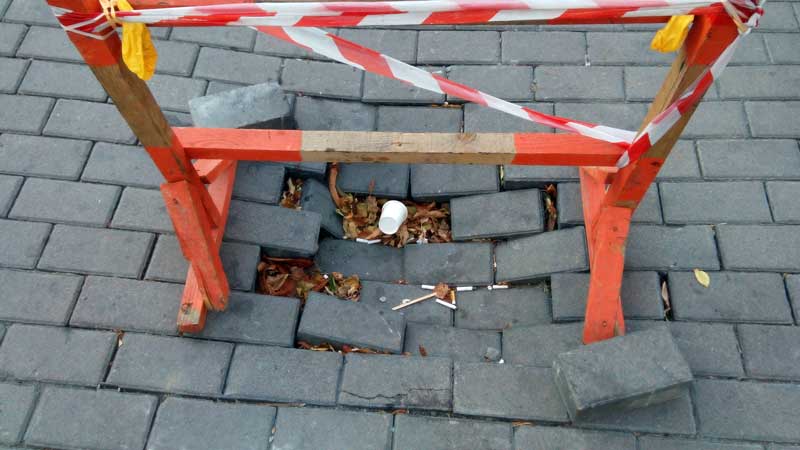 Wooden safety barrier on top of hole where concrete patio pavers have sunk into the ground and come apart.