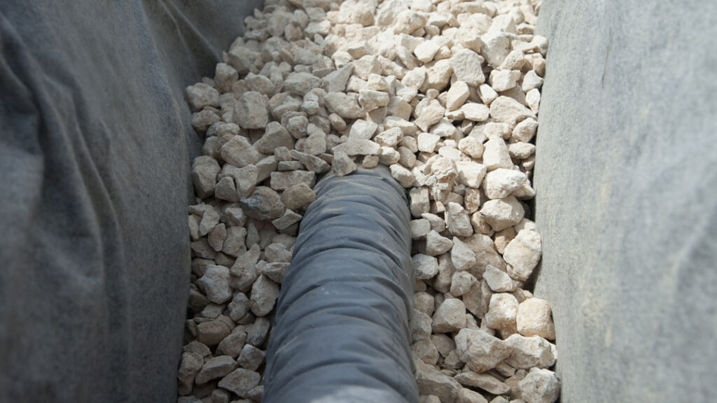 French drain tubing that is surrounded by rocks in the trench. The tubing will be covered to complete the french drain.